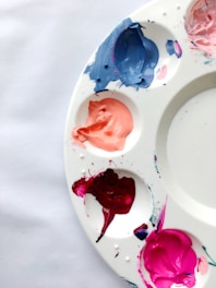 A white artist's palette holds various colors of paint, including shades of blue, peach, red, and pink. The paints are arranged in individual wells on the palette, some smeared slightly beyond the wells.
