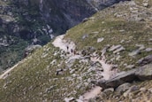 A rugged mountain trail winds through a rocky landscape, flanked by patches of green vegetation. Several hikers wearing backpacks are making their way along the path, indicating a trekking or hiking activity. The terrain is uneven and composed of large boulders and grass-covered slopes.