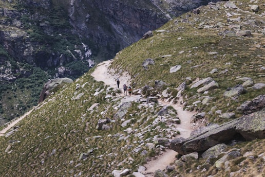 A rugged mountain trail winds through a rocky landscape, flanked by patches of green vegetation. Several hikers wearing backpacks are making their way along the path, indicating a trekking or hiking activity. The terrain is uneven and composed of large boulders and grass-covered slopes.