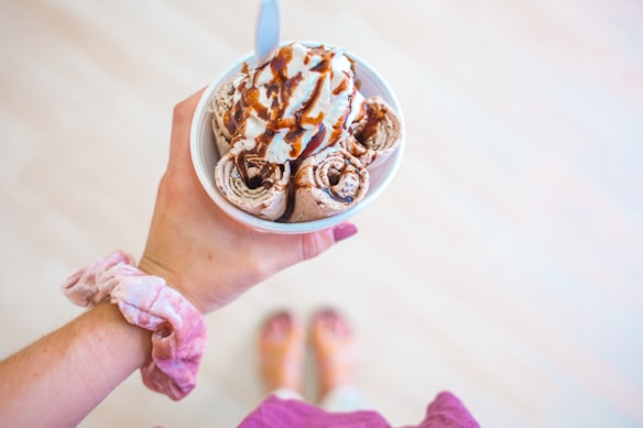 A person holds a cup containing rolled ice cream topped with whipped cream and chocolate syrup. The hand holding the cup has a pink scrunchie on the wrist. Gentle colors and a soft focus give the scene a casual, summery feel.