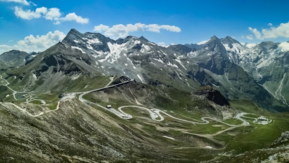 A breathtaking mountain range with snow-capped peaks under a clear blue sky. Winding roads are visible, carving through the lush green valleys and rocky terrain. The landscape is expansive, showcasing the grandeur of the natural scenery.