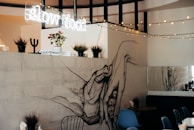 An interior space featuring a large, artistic hand mural on a wall, with potted plants and a 'slow food' neon sign above. String lights are draped across, adding a cozy ambiance. There is modern furniture including blue chairs and a table, enhancing the contemporary aesthetic.
