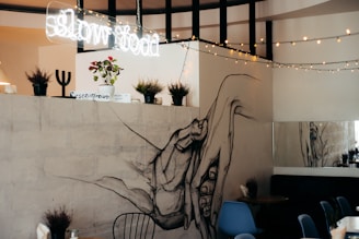 An interior space featuring a large, artistic hand mural on a wall, with potted plants and a 'slow food' neon sign above. String lights are draped across, adding a cozy ambiance. There is modern furniture including blue chairs and a table, enhancing the contemporary aesthetic.
