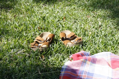 A pair of brown sandals resting on lush green grass, with a checkered red and blue cloth partially visible beside them.