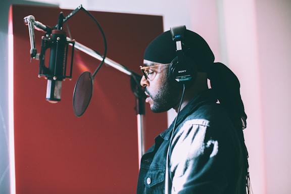 A person wearing headphones stands in front of a microphone in a recording studio. The individual appears to be focused and is singing or speaking into the microphone. The background shows acoustic panels and studio equipment, with lighting providing a soft ambiance.