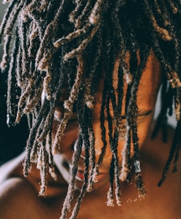 A close-up view of a person with their head slightly bowed, showcasing their long, textured dreadlocks that cover most of their face. The lighting highlights the texture and detail of the hair while casting shadows on their skin.