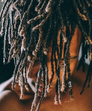 A close-up view of a person with their head slightly bowed, showcasing their long, textured dreadlocks that cover most of their face. The lighting highlights the texture and detail of the hair while casting shadows on their skin.