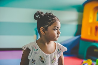 A young child with a serious expression is sitting indoors. The child has dark hair tied in a bun and is wearing a white shirt with colorful floral embroidery. The background features a play area with colorful equipment, including a plastic playhouse in shades of blue, orange, and yellow.