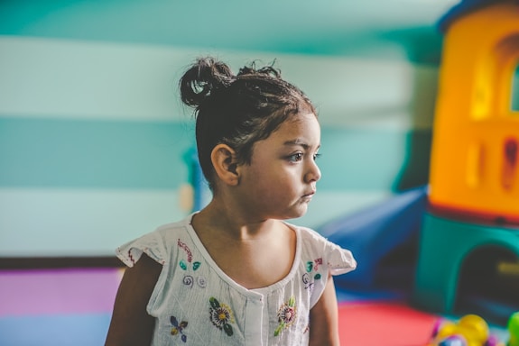 A young child with a serious expression is sitting indoors. The child has dark hair tied in a bun and is wearing a white shirt with colorful floral embroidery. The background features a play area with colorful equipment, including a plastic playhouse in shades of blue, orange, and yellow.