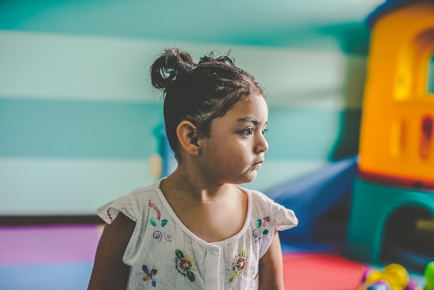 A young child with a serious expression is sitting indoors. The child has dark hair tied in a bun and is wearing a white shirt with colorful floral embroidery. The background features a play area with colorful equipment, including a plastic playhouse in shades of blue, orange, and yellow.