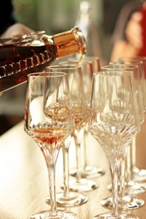 A row of elegant, clear crystal glasses being filled with a golden-brown liquid from a dark glass bottle. The scene is well-lit with a soft, warm ambiance, highlighting the intricate detailing of the glassware.