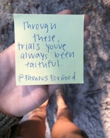 A person is holding a yellow sticky note with handwritten text that reads 'Through these trials you've always been faithful. @FamousForGood'. The background features a blurry view of a sandy or gravel path, and the person wearing shorts and shoes is partially visible.