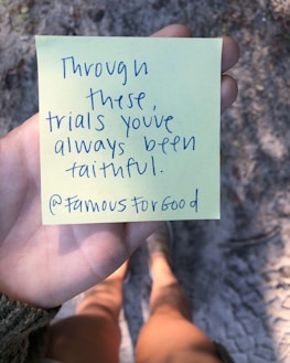 A person is holding a yellow sticky note with handwritten text that reads 'Through these trials you've always been faithful. @FamousForGood'. The background features a blurry view of a sandy or gravel path, and the person wearing shorts and shoes is partially visible.