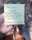 A person is holding a yellow sticky note with handwritten text that reads 'Through these trials you've always been faithful. @FamousForGood'. The background features a blurry view of a sandy or gravel path, and the person wearing shorts and shoes is partially visible.