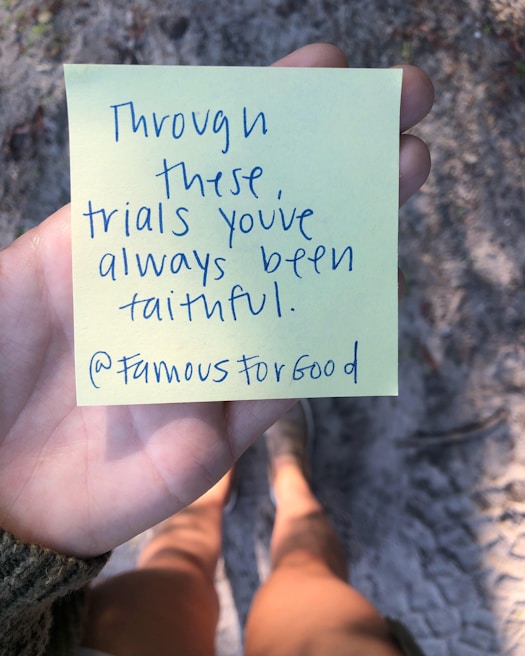 A person is holding a yellow sticky note with handwritten text that reads 'Through these trials you've always been faithful. @FamousForGood'. The background features a blurry view of a sandy or gravel path, and the person wearing shorts and shoes is partially visible.