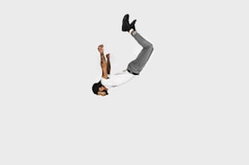 A person is captured mid-air in a flipped position against a plain white background. They are wearing a white t-shirt, gray pants, and black shoes, with tattoos visible on their arms. The minimalistic backdrop emphasizes the dynamic movement and the acrobatic pose.