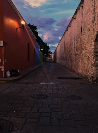 A narrow cobblestone alley is flanked by a vibrant orange wall on one side and an aged, weathered stone wall on the other. Street lamps cast a warm glow as the evening sky transitions from blue to pink and purple hues, with scattered clouds adding texture.