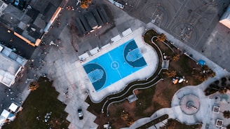 An aerial view of a brightly colored blue basketball court surrounded by concrete pathways, grassy areas, and scattered people. Buildings and other basketball courts are visible on the outer edges, giving a sense of a bustling urban park.