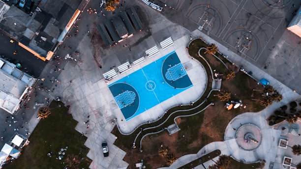 An aerial view of a brightly colored blue basketball court surrounded by concrete pathways, grassy areas, and scattered people. Buildings and other basketball courts are visible on the outer edges, giving a sense of a bustling urban park.