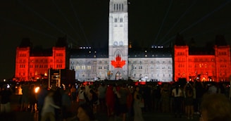 A large building is illuminated at night with light projections. The central tower is lit in white with a large red maple leaf displayed on it. The surrounding sections of the building are illuminated in red, creating a striking contrast against the night sky. Crowds of people are gathered in the foreground, observing the light show.