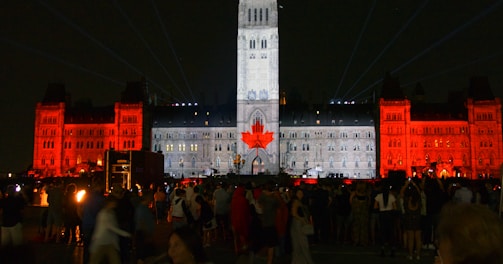 A large building is illuminated at night with light projections. The central tower is lit in white with a large red maple leaf displayed on it. The surrounding sections of the building are illuminated in red, creating a striking contrast against the night sky. Crowds of people are gathered in the foreground, observing the light show.