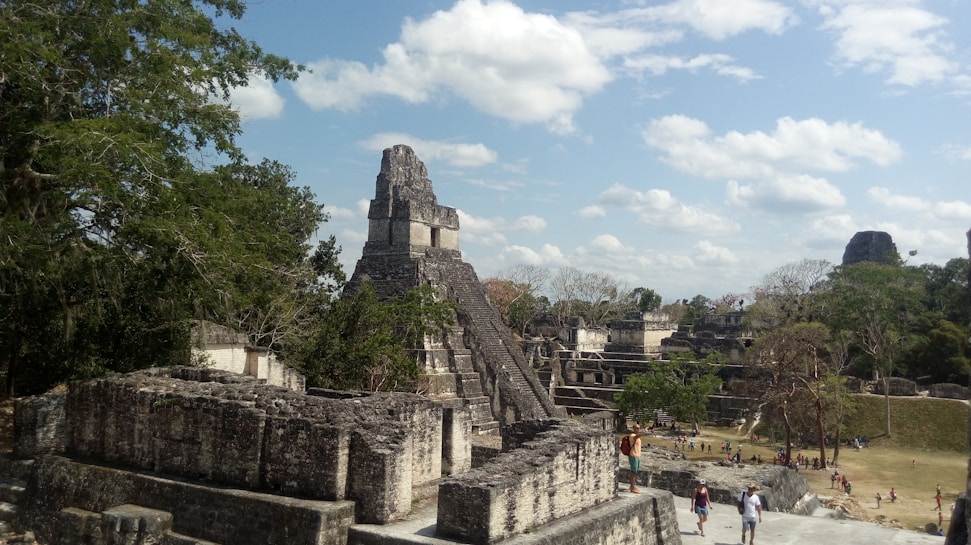 Ancient stone ruins with a large, stepped pyramid dominate the landscape, surrounded by scattered trees and a few clouds in the sky. Tourists are walking around the ruins, giving a sense of scale and human presence.