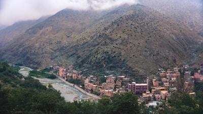 A picturesque village nestled in a mountainous region with a river running alongside. The houses are built closely together, displaying a mix of earthy tones and vibrant colors. The mountains in the backdrop are lush with patches of greenery and partially shrouded in mist, giving a serene and tranquil atmosphere.