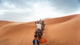 A group of people riding camels traverses expansive sand dunes under a partly cloudy sky. The line of camels stretches into the distance, climbing a ridge of ochre-colored sand.