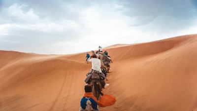 A group of people riding camels traverses expansive sand dunes under a partly cloudy sky. The line of camels stretches into the distance, climbing a ridge of ochre-colored sand.