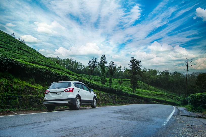A white car is parked on the side of an empty road that curves through lush green tea plantations. The sky is clear and vibrant with scattered white clouds, adding a sense of openness. Tall trees line the horizon, and electricity poles can be seen along the road, indicating a rural setting.