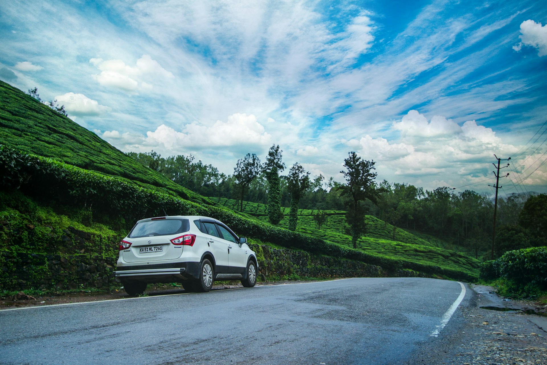A white cab cruising along a scenic road bordered by lush green tea gardens of Assam under a bright blue sky.