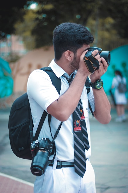 A person in a white school uniform is holding a camera to their face, appearing to take a photograph. They are wearing a black backpack and have a second camera hanging around their neck. The background is slightly blurred with some greenery and a hint of buildings.