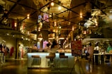 A modern, dimly lit cafe or lounge area with stylish hanging light bulbs reflecting off a mirrored ceiling. A woman is seated at a bar with two stools, near a sign that reads 'Hello It's Choux.' The area is part of a larger retail space with mannequins displaying clothing in the background.