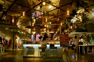 A modern, dimly lit cafe or lounge area with stylish hanging light bulbs reflecting off a mirrored ceiling. A woman is seated at a bar with two stools, near a sign that reads 'Hello It's Choux.' The area is part of a larger retail space with mannequins displaying clothing in the background.
