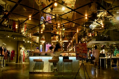 A modern, dimly lit cafe or lounge area with stylish hanging light bulbs reflecting off a mirrored ceiling. A woman is seated at a bar with two stools, near a sign that reads 'Hello It's Choux.' The area is part of a larger retail space with mannequins displaying clothing in the background.