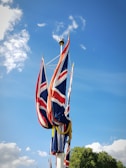 Multiple flags featuring the Union Jack design are attached to a pole, waving against a clear blue sky with scattered clouds. A patch of greenery is visible at the bottom right.
