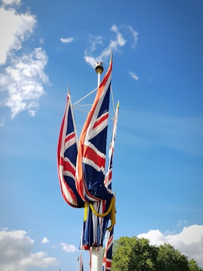 Multiple flags featuring the Union Jack design are attached to a pole, waving against a clear blue sky with scattered clouds. A patch of greenery is visible at the bottom right.