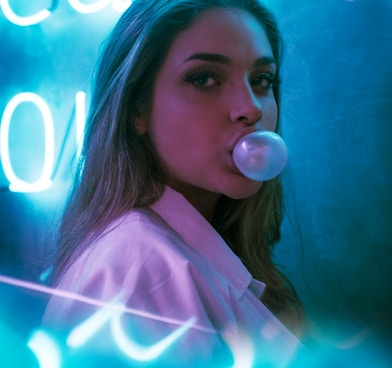 selective focus photo of woman blowing gum standing in front of turned-on neon signage
