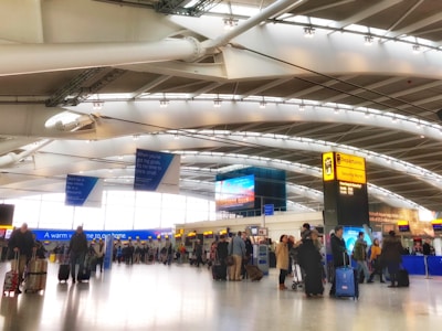 A busy airport terminal with passengers walking and standing near check-in counters. The interior features high, curved ceilings and prominent signage for departures and security. Several carry-on suitcases and travel bags are visible.