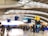 A busy airport terminal with passengers walking and standing near check-in counters. The interior features high, curved ceilings and prominent signage for departures and security. Several carry-on suitcases and travel bags are visible.