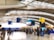 A busy airport terminal with passengers walking and standing near check-in counters. The interior features high, curved ceilings and prominent signage for departures and security. Several carry-on suitcases and travel bags are visible.