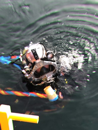 A diver wearing a heavy-duty diving helmet is submerged in dark water, creating ripples around. The helmet is metallic with various attachments and a transparent front panel. Brightly colored cables are seen near the diver, enhancing the underwater setting.
