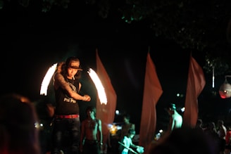 A person is performing a fire dance at night, illuminated by flames creating arcs around them. They are wearing a hat and a sleeveless shirt with visible tattoos. Other individuals and large, red fabric flags are visible in the background, contributing to a dynamic and lively atmosphere.