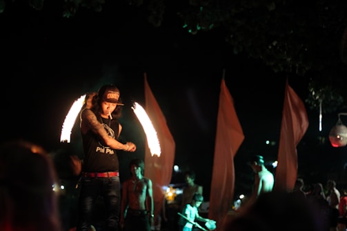 A person is performing a fire dance at night, illuminated by flames creating arcs around them. They are wearing a hat and a sleeveless shirt with visible tattoos. Other individuals and large, red fabric flags are visible in the background, contributing to a dynamic and lively atmosphere.