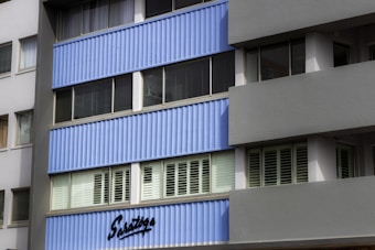 The image shows a section of a modern building, featuring blue corrugated panels and multiple glass windows with shutters. The fa&ccedil;ade is mostly uniform with clean lines, and there is a sign that reads 'Saratoga' near the bottom. The structure has a contemporary architectural style.