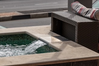 A small water feature with a gentle stream of water cascading into a built-in stone pool. The pool area is tiled with green mosaic tiles, and beside it is a wicker bench with a striped cushion resting on top. The background shows a blurred view of a street with a curb.