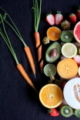 A collection of fresh fruits and vegetables is laid out on a dark textured surface. Visible are whole and sliced oranges, strawberries, kiwis, and an avocado along with several small carrots with their greens still attached. There is also a slice of lemon, lime, melon, and a persimmon.