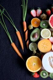 A collection of fresh fruits and vegetables is laid out on a dark textured surface. Visible are whole and sliced oranges, strawberries, kiwis, and an avocado along with several small carrots with their greens still attached. There is also a slice of lemon, lime, melon, and a persimmon.