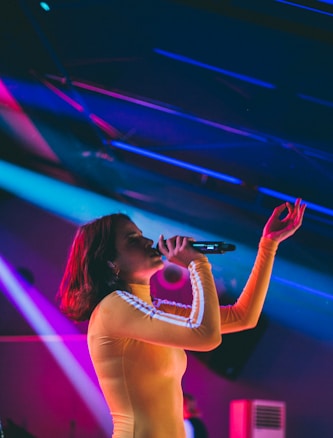 A person in a yellow outfit is singing on stage, holding a microphone, with colorful lights and a dynamic background enhancing the lively atmosphere.