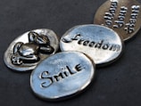 Four silver metallic tokens lie on a dark gray surface. Each token has a word or an image engraved on it. One token has a symbol of a turtle, another says 'Freedom', the third reads 'Smile', and the fourth one says 'Follow Your Heart'. The tokens have a shiny, polished appearance with a slightly worn texture.